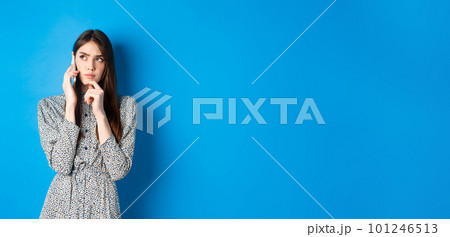 Pensive woman talking on phone and thinking, looking aside thoughtful, standing against blue background 101246513