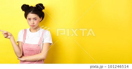 Sad and gloomy asian girl with glamour makeup, frowning unfair and pointing left at logo, looking disappointed, standing on yellow background 101246514