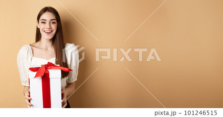 Happy valentines day. Cheerful smiling girl giving surprise gift to lover, looking at camera and holding present in box, standing on beige background 101246515