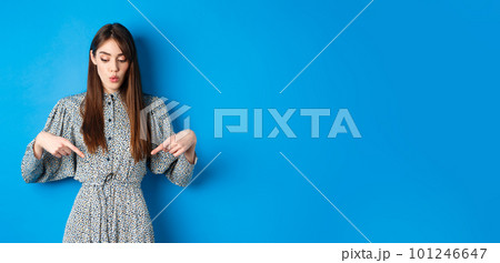 Intrigued beautiful girl in dress say wow, looking and pointing down at advertisement, showing promo with interested face, blue background 101246647