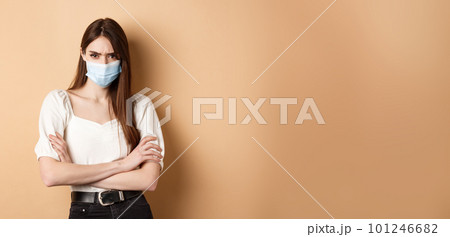 Covid-19 and lifestyle concept. Angry woman look with despise, wearing medical mask, cross arms on chest and stare with condemn, standing on beige background 101246682