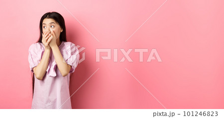 Shocked and startled asian woman looking aside at logo, covering mouth with hands speechless, standing in dress on pink background 101246823