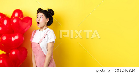 Valentines day and relationship concept. Fashion girl looking amazed, gasping and say wow, staring left while standing near red hearts balloons, surprise from lover, yellow background 101246824