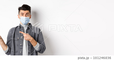 Pandemic lifestyle, healthcare and medicine concept. Young guy in face mask inviting look here, pointing fingers left at logo, standing on white background 101246836