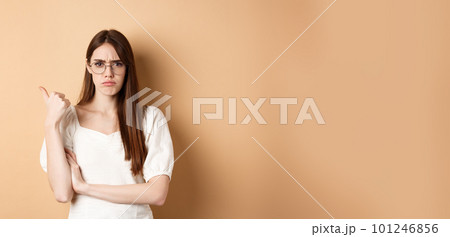 Disappointed girl in glasses frowning, pointing aside at bad product, disapprove and dislike something, standing on beige background 101246856