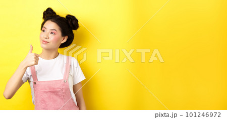 Glamour asian girl with pink makeup and summer clothes, looking left at logo with thumb up, approve and praise, standing on yellow background 101246972