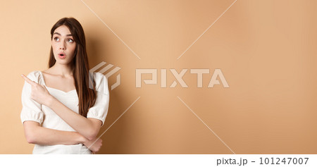 Young woman in dress looking and pointing at upper left corner wondered, saying wow, checking out advertisement with intrigued face, beige background 101247007