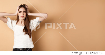 Frustrated young woman holding hands on head and feeling sad, standing distressed and troubled on beige background Frustrated young woman holding hands on head and feeling sad, standing distressed and troubled on beige background 101247155