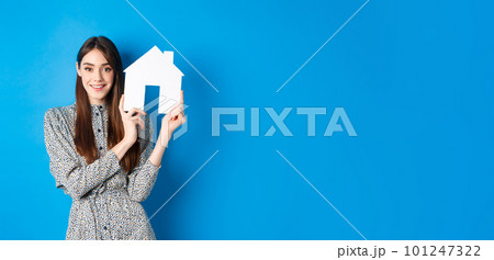 Real estate, realtors and insurance concept. Beautiful lady in dress showing paper house cutout, suggest property for sale, standing on blue background 101247322