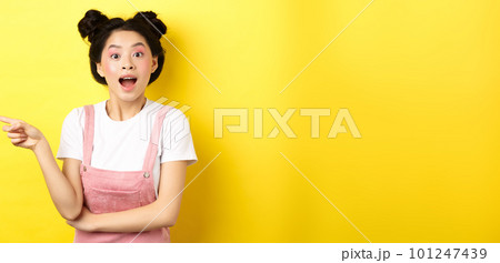 Excited asian teen girl pointing finger left at logo and looking happy, showing advertisement, standing on yellow background 101247439