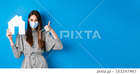 Real estate, covid-19 and pandemic concept. Candid woman in medical mask showing thumb up and paper house cutout, reanting apartment, standing on blue background Real estate, covid-19 and pandemic concept. Candid woman in medical mask showing thumb up and paper house cutout, reanting apartment, standing on blue background 101247467