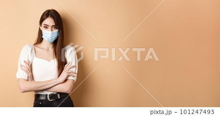 Covid-19 and lifestyle concept. Arrogant woman in medical mask frowning, cross arms on chest and looking confident at camera, beige background 101247493