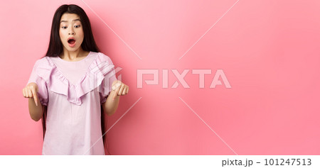 Surprised asian woman in awe, drop jaw and gasping, pointing fingers down at amazing promo offer, standing impressed against pink background Surprised asian woman in awe, drop jaw and gasping, pointing fingers down at amazing promo offer, standing impressed against pink background 101247513