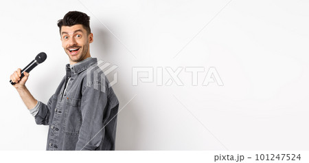 Profile of handsome smiling man holding mic, turn head at camera with excited face, singing karaoke and perform standup, standing on white background 101247524