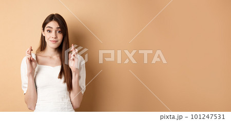 Hopeful tender girl in white blouse making wish, holding fingers crossed and smiling, praying for good luck, waiting for results, beige background Hopeful tender girl in white blouse making wish, holding fingers crossed and smiling, praying for good luck, waiting for results, beige background 101247531