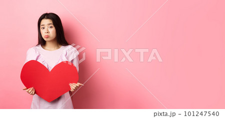 Valentines concept. Single teenage asian girl wants to fall in love, looking sad and lonely at camera, sulking distressed on lovers day, holding big red heart cutout, pink background Valentines concept. Single teenage asian girl wants to fall in love, looking sad and lonely at camera, sulking distressed on lovers day, holding big red heart cutout, pink background 101247540