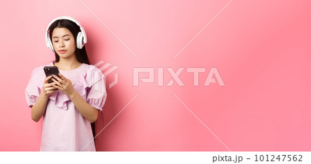 Young girl listening music in headphones and chatting on mobile phone, standing in dress against pink background Young girl listening music in headphones and chatting on mobile phone, standing in dress against pink background 101247562