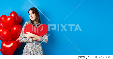 Valentines day. Beautiful and romantic woman looking pensive at balloons, hugging big red heart and smiling, waiting for love, blue background 101247568