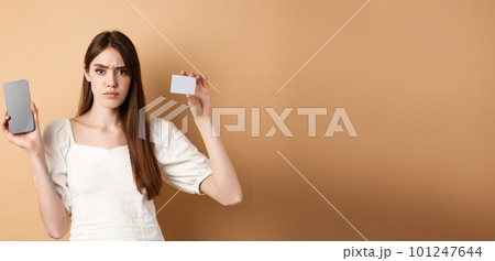 Sad and disappointed girl showing empty smartphone screen and plastic credit card, complaining and frowning, standing on beige background 101247644