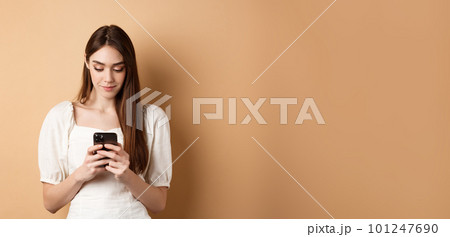 Pretty woman texting on smartphone cell phone, reading screen and smiling, standing on beige background 101247690