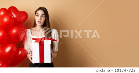 Valentines day. Pensive cute girl guessing who made her gift, holding present and looking curious at upper left corner, standing near romantic heart balloons, beige background 101247728