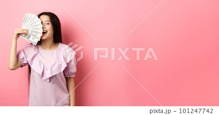 Happy rich asian woman showing money and smiling, shopping with cash, standing in dress against pink background Happy rich asian woman showing money and smiling, shopping with cash, standing in dress against pink background 101247742