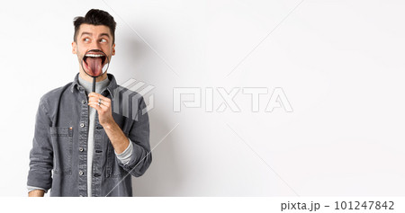 Handsome positive guy showing white perfect teeth and tongue with magnifying glass, looking left at logo, standing against white background Handsome positive guy showing white perfect teeth and tongue with magnifying glass, looking left at logo, standing against white background 101247842