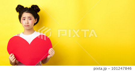 Heartbroken asian woman showing big red heart cutout and looking sad, feeling lonely on lovers day, showing valentine card at camera, yellow background 101247846