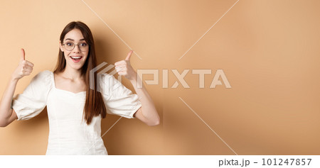 Excited young woman in glasses showing thumbs up and smiling, praising good optic store, approve and like eyewear, standing on beige background 101247857