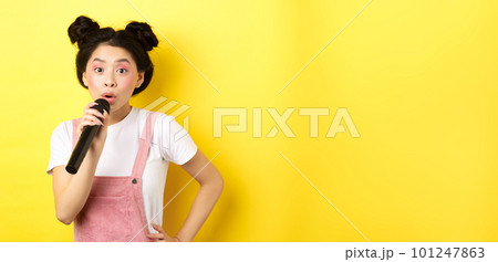Cute asian teen girl with bright makeup, singing in microphone karaoke, standing against yellow background Cute asian teen girl with bright makeup, singing in microphone karaoke, standing against yellow background 101247863