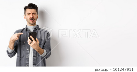 Sad crying guy pointing at smartphone and sobbing, complaining or feeling miserable, standing on white background 101247911