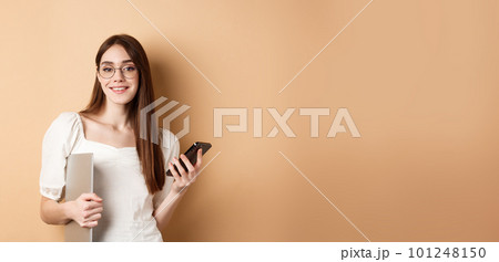 Elegant working woman in glasses holding laptop computer and smartphone, smiling pleased at camera, standing on beige background 101248150