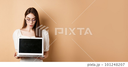 Excited girl in glasses show laptop screen, demonstrate online offer on computer, standing against beige background Excited girl in glasses show laptop screen, demonstrate online offer on computer, standing against beige background 101248207
