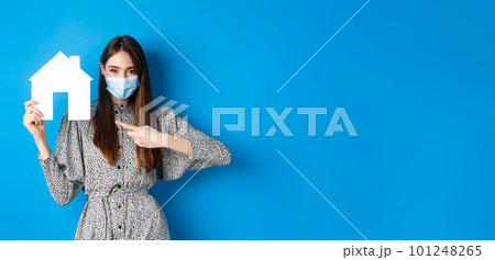 Real estate, covid-19 and pandemic concept. Smiling female model in medical mask pointing at paper house cutout, standing on blue background 101248265