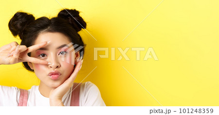 Close-up of cute asian woman with bright glamour makeup, long fingernails, showing v-sign and pouting silly at camera, standing kawaii on yellow background 101248359