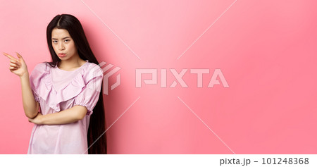 Sad and grumpy asian girl sulking, pointing left and look at camera jealous, want something, complaining and showing desired thing, standing on pink background 101248368