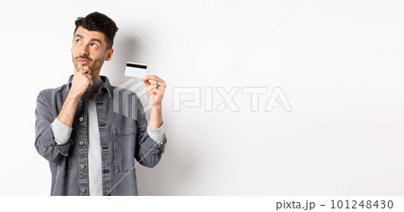 Thoughtful guy looking at upper left corner logo and holding plastic credit card, thinking about shopping, standing on white background Thoughtful guy looking at upper left corner logo and holding plastic credit card, thinking about shopping, standing on white background 101248430