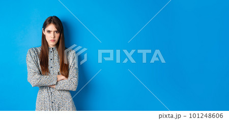 Angry frowning woman in dress, cross arms on chest and feel offended about something unfair, standing upset against blue background 101248606