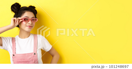 Beautiful asian girl put on stylish sunglasses and smiling, wearing trendy summer outfit, standing on yellow background 101248697