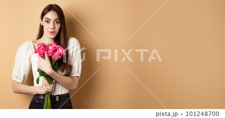 Valentines day. Surprised tender girl thanking for flowers and smiling, holding red roses and smiling grateful, standing on beige background Valentines day. Surprised tender girl thanking for flowers and smiling, holding red roses and smiling grateful, standing on beige background 101248700