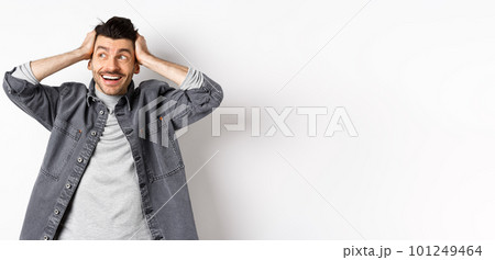 Excited young man looking relieved and happy aside, smiling and staring at logo, holding hands on head cheerful, standing on white background Excited young man looking relieved and happy aside, smiling and staring at logo, holding hands on head cheerful, standing on white background 101249464
