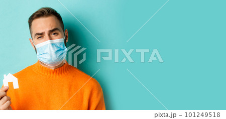 Real estate and coronavirus pandemic concept. Close-up of sad man in face mask showing face mask and grimacing upset, standing over light blue background 101249518