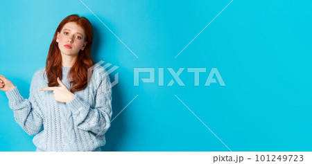 Moody teenage girl with red hair, pointing fingers left at logo, staring bothered and bored, standing over blue background Moody teenage girl with red hair, pointing fingers left at logo, staring bothered and bored, standing over blue background 101249723