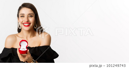 Fashion and beauty concept. Close-up of gorgeous brunette woman with red lips, black dress, showing engagement ring in box and smiling, getting married, standing over white background Fashion and beauty concept. Close-up of gorgeous brunette woman with red lips, black dress, showing engagement ring in box and smiling, getting married, standing over white background 101249743