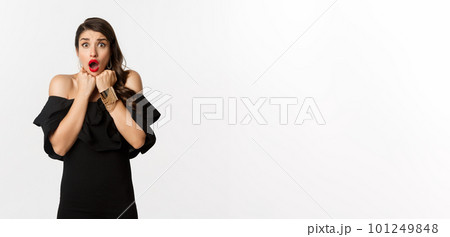 Portrait of glamour woman looking scared and shocked at camera, staring at something with fear, standing in black dress against white background 101249848