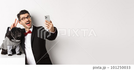Cheerful dog owner in suit celebrating New Year with dog, taking selfie on smartphone near cute black pug in costume, white background Cheerful dog owner in suit celebrating New Year with dog, taking selfie on smartphone near cute black pug in costume, white background 101250033