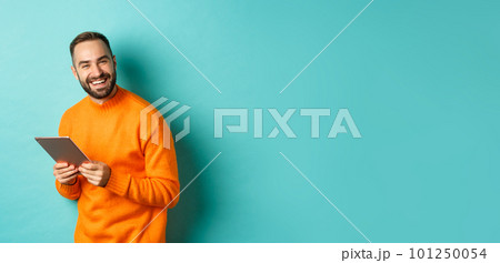 Handsome bearded man using digital tablet, laughing at camera, standing happy against light blue background 101250054