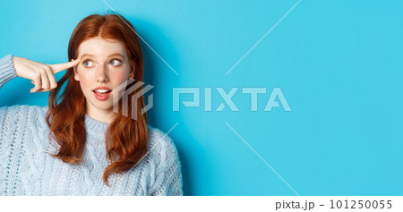 Close-up of cute redhead girl searching for solution, pointing at head and looking left at logo, standing over blue background Close-up of cute redhead girl searching for solution, pointing at head and looking left at logo, standing over blue background 101250055