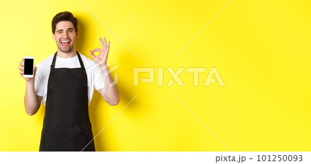 Handsome coffee shop worker showing ok sign and smartphone screen, recommending application, standing over yellow background Handsome coffee shop worker showing ok sign and smartphone screen, recommending application, standing over yellow background 101250093