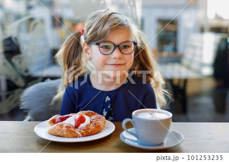 Adorable smiling girl with glasses have a breakfast in a cafe. Preschool child with glasses drinking chocolate and eating bakery pastry croissant or cake. Happy children, healthy food and meal. 101253235